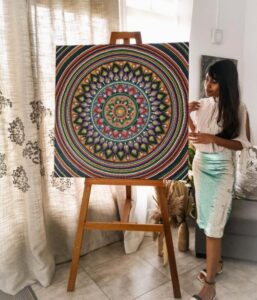Atasha Ramanah - Intricate Mandala Artwork on Display A colorful mandala painting with intricate patterns displayed on an easel, with Atasha standing to the side.