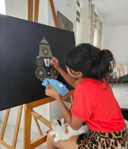 Atasha - Young Artist Creates Ganesh Mandala Masterpiece Atasha painting Ganesh on black canvas with cat beside her.
